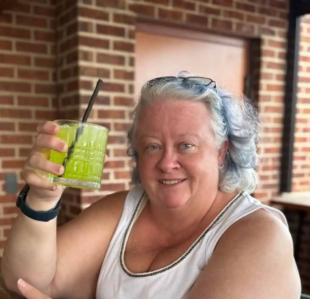 A woman with light blue-gray hair holds up a glass of green drink with a straw, sitting outside by a brick wall, smiling at the camera.