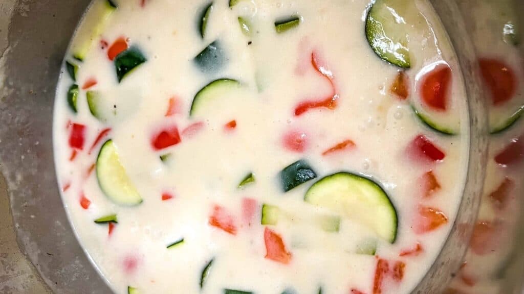 Bowl of thick, creamy batter mixed with chopped vegetables including zucchini and red bell pepper.