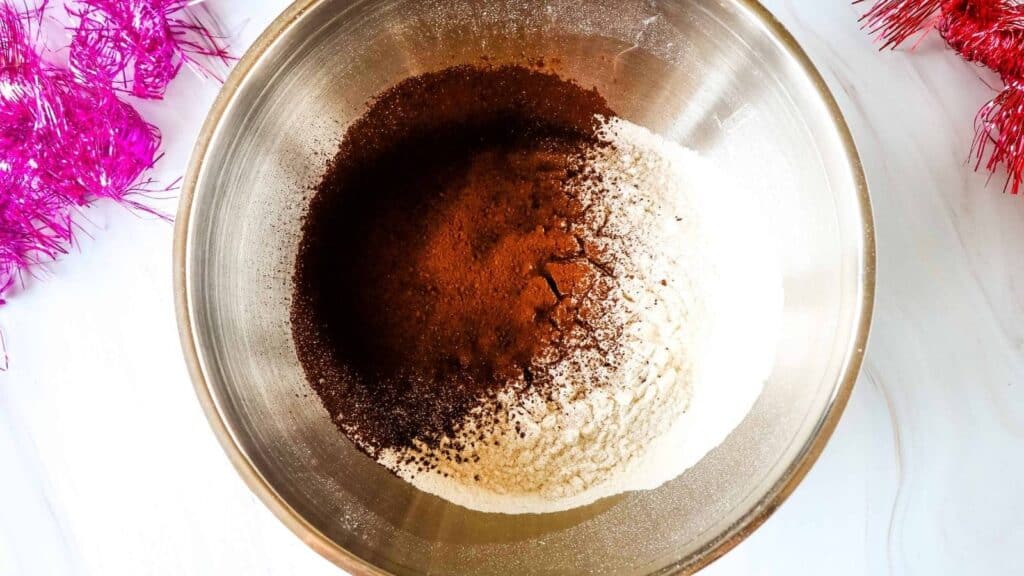 A metal bowl containing unmixed dry ingredients including flour, cocoa powder, and possibly baking powder, on a white surface with pink tinsel nearby.