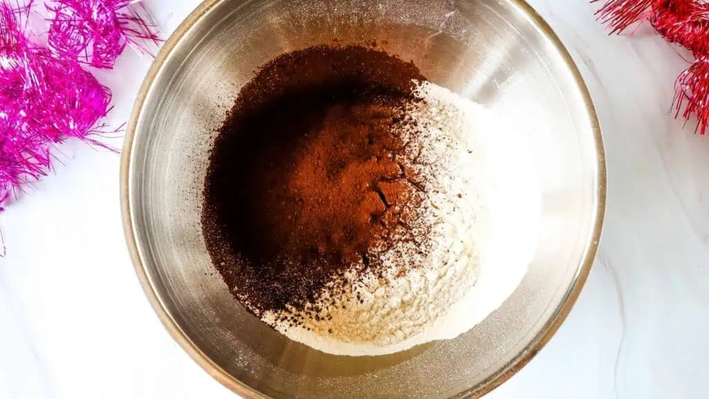 A metal bowl containing unmixed dry ingredients including flour, cocoa powder, and possibly baking powder, on a white surface with pink tinsel nearby.