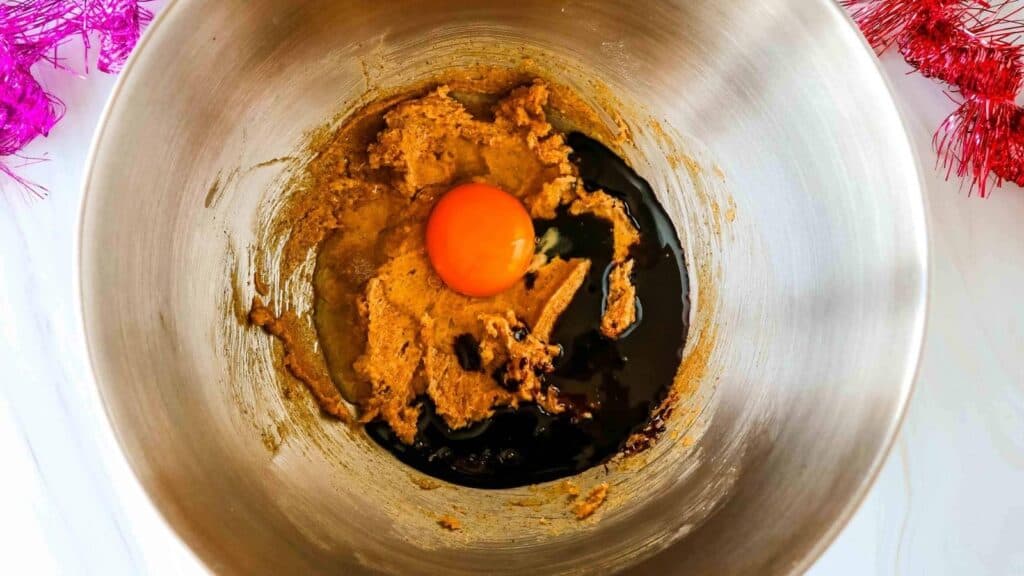 A metal mixing bowl containing brown sugar mixture, a cracked raw egg, and dark liquid ingredients ready to be mixed.