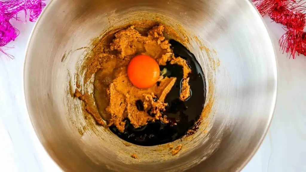 A metal mixing bowl containing brown sugar mixture, a cracked raw egg, and dark liquid ingredients ready to be mixed.