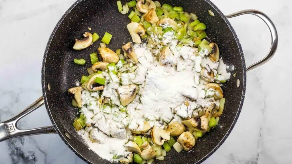 Chopped celery, mushrooms, and flour cooking in a black skillet on a white marble countertop.