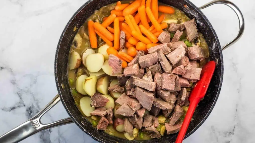 A skillet filled with chopped beef, halved baby potatoes, baby carrots, and a savory broth, with a red spatula resting inside the pan.