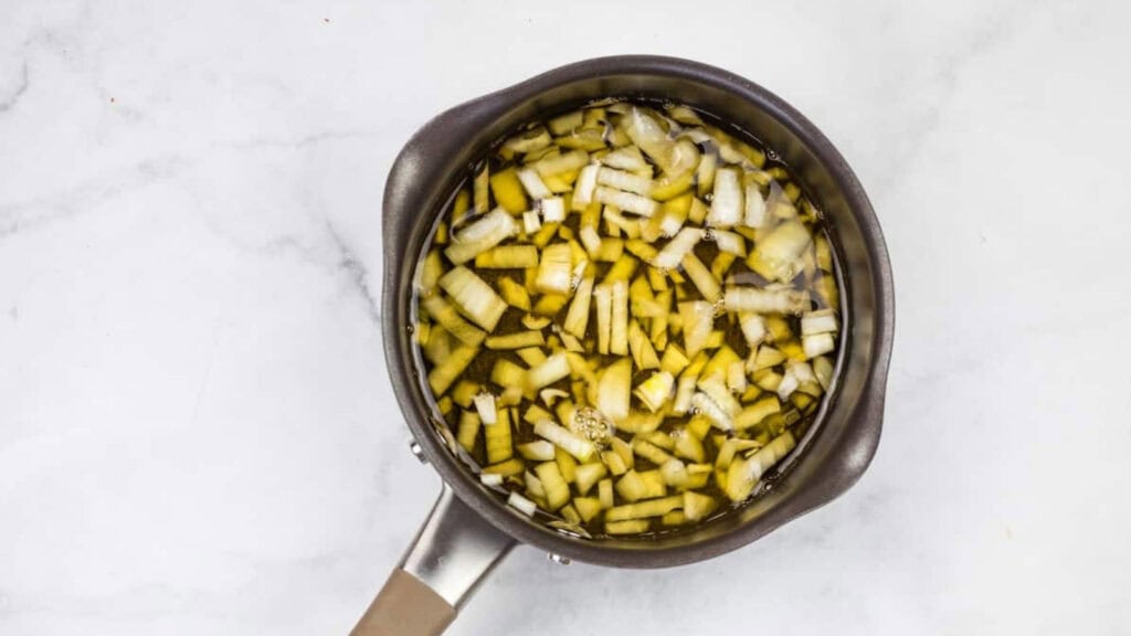 A saucepan filled with chopped onions and liquid sits on a white marble surface.