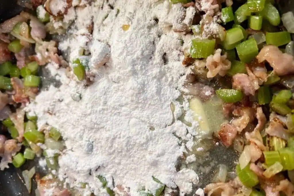 Chopped celery, onions, and bacon are being sautéed in a pan with a mound of flour on top, ready to be mixed in.