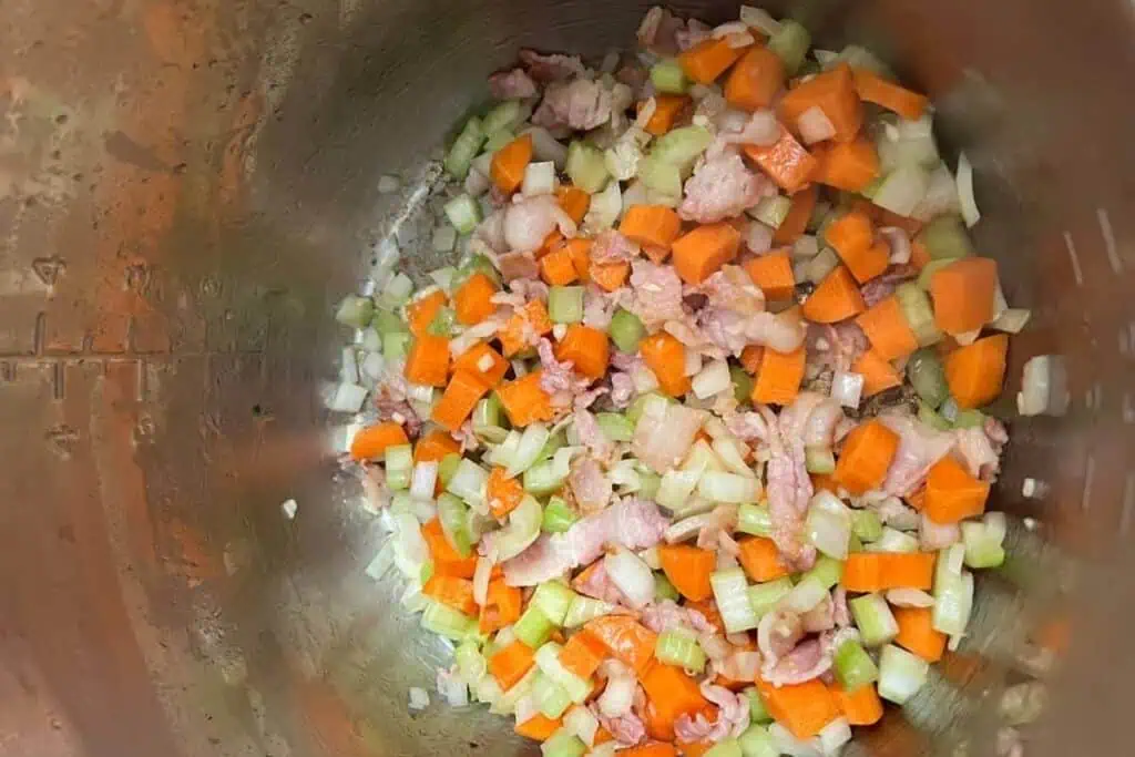 Chopped carrots, celery, onions, and pieces of bacon are being sautéed in a metal pot.