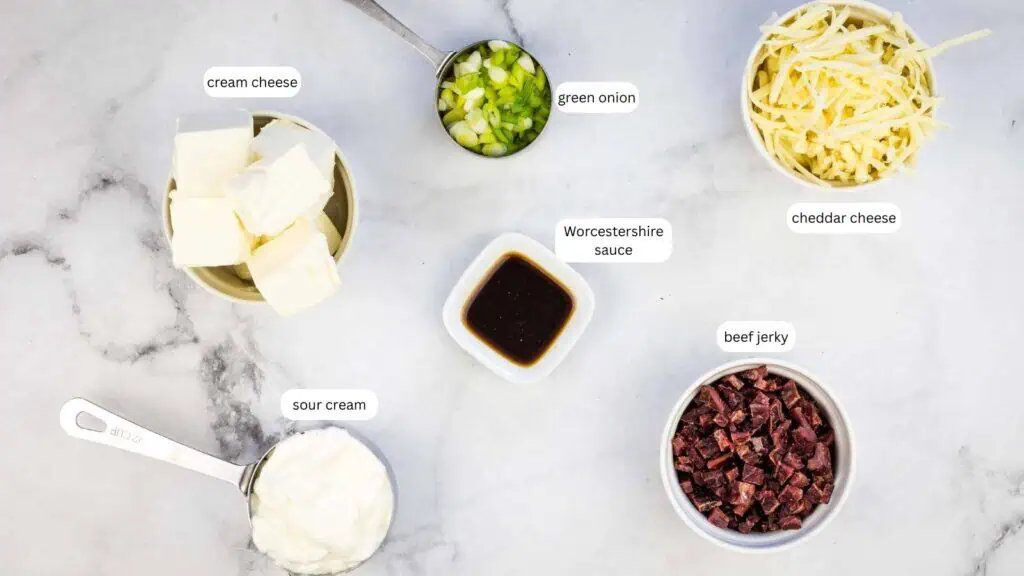 Ingredients on a marble surface: cream cheese, green onion, cheddar cheese, Worcestershire sauce, sour cream, and chopped beef jerky, each in separate bowls or measuring cups.