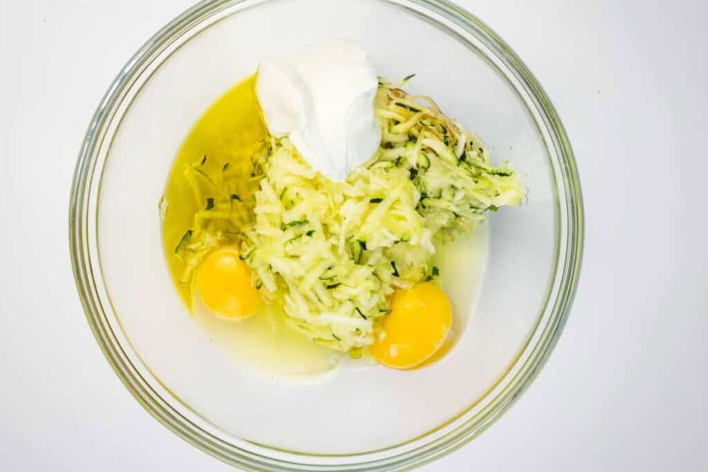 A glass bowl containing two raw eggs, shredded zucchini, and a dollop of sour cream, viewed from above on a white background.