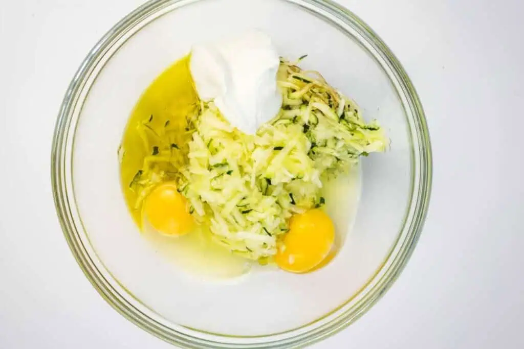 A glass bowl containing two raw eggs, shredded zucchini, and a dollop of sour cream, viewed from above on a white background.