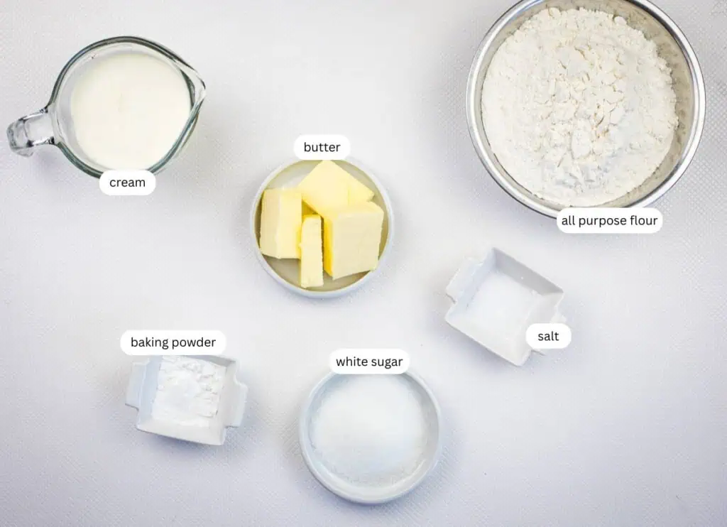 Top-down view of labeled bowls containing cream, butter, all purpose flour, baking powder, white sugar, and salt on a white surface.