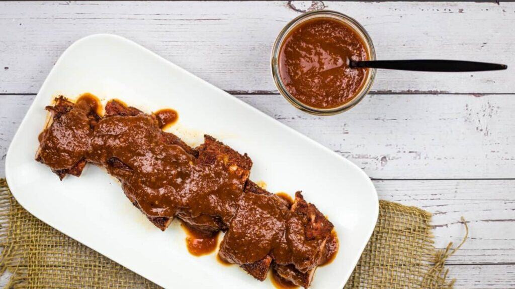 A rectangular white plate with pieces of meat covered in brown sauce, next to a jar of the same sauce with a black spoon, on a rustic wooden surface.