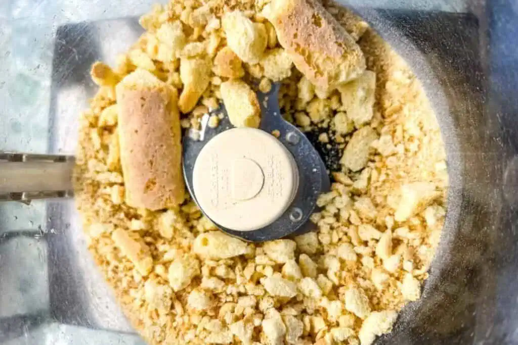 Crushed ladyfinger cookies inside a food processor bowl with stainless steel blades.