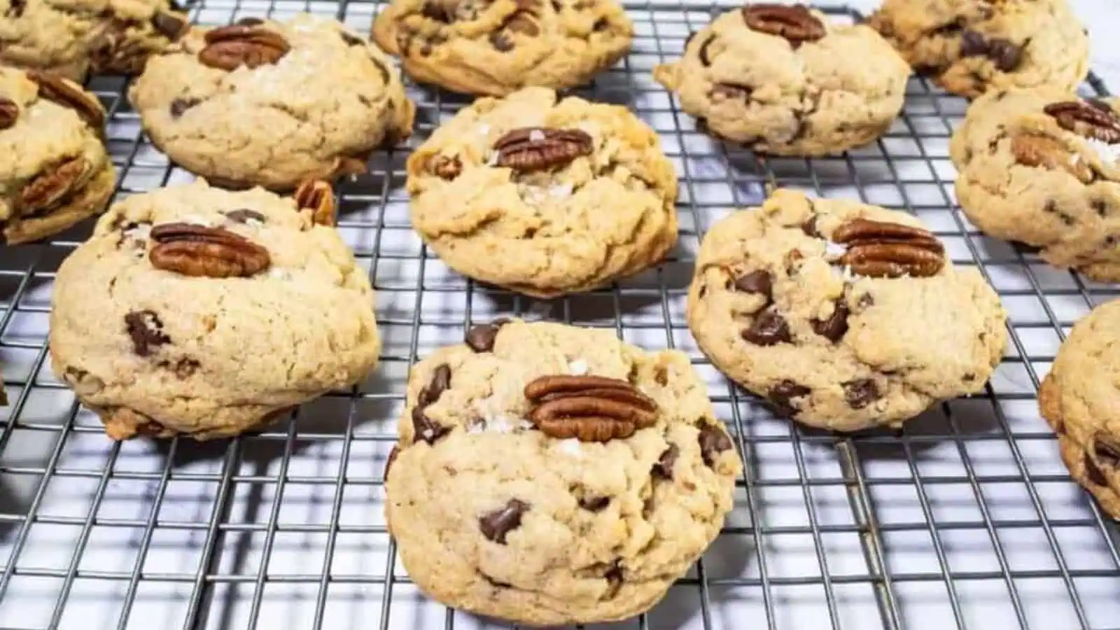 Chocolate chip cookies with pecan halves on top are cooling on a metal wire rack.