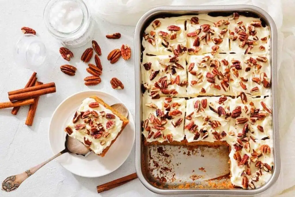 A pan of frosted sheet cake topped with chopped pecans, with one slice served on a plate beside cinnamon sticks and whole pecans.