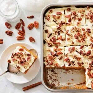 A pan of carrot cake bars topped with chopped pecans, with one slice served on a plate beside cinnamon sticks and whole pecans.