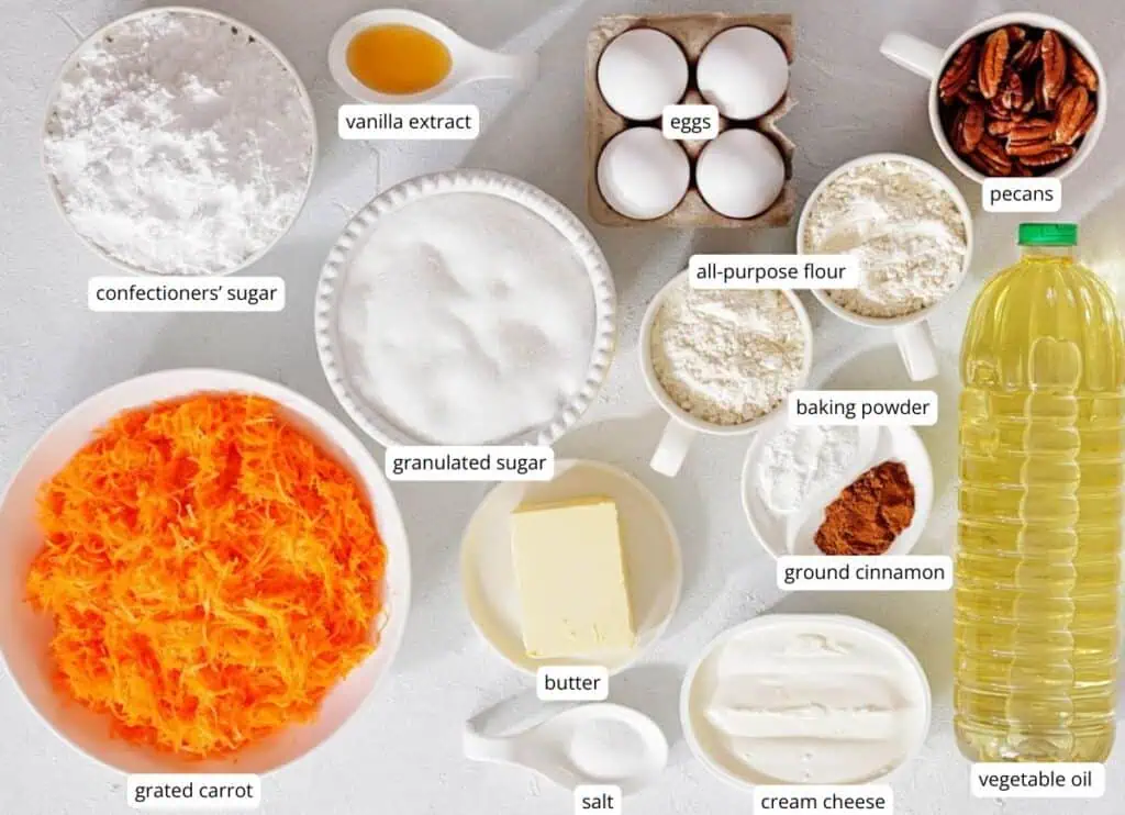 Assorted labeled ingredients for baking, including grated carrot, sugars, eggs, butter, flour, baking powder, ground cinnamon, salt, cream cheese, vanilla extract, pecans, and vegetable oil.