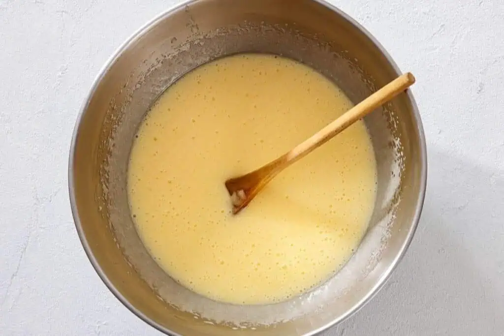 A metal mixing bowl with pale yellow batter and a wooden spoon resting inside, placed on a light-colored surface.
