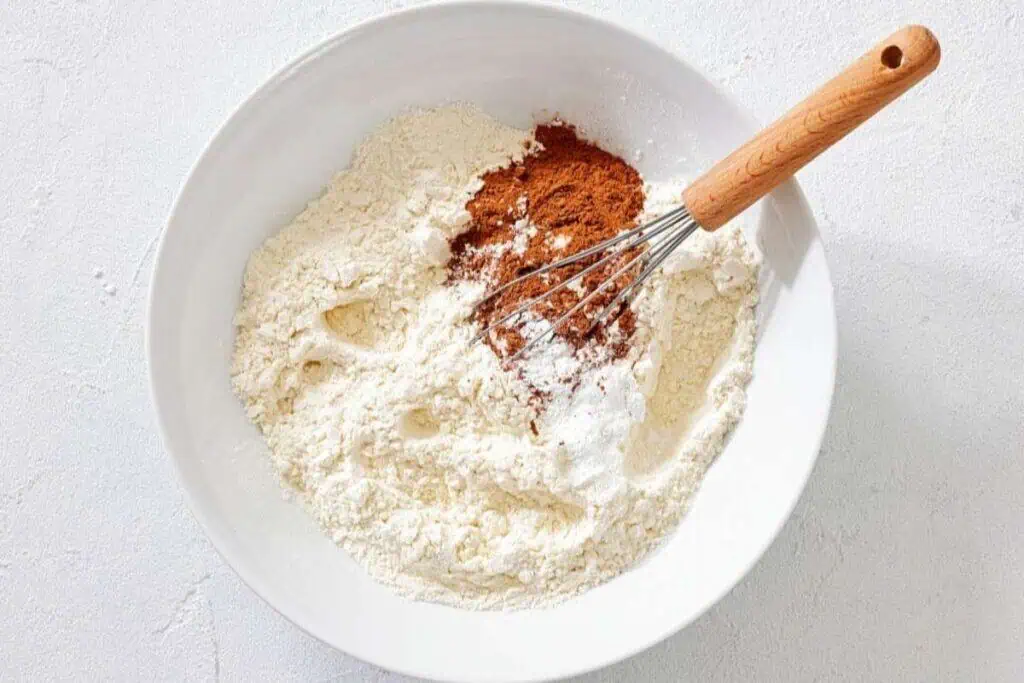 A white bowl with flour, cinnamon, baking powder, and other dry ingredients being whisked together on a light surface.