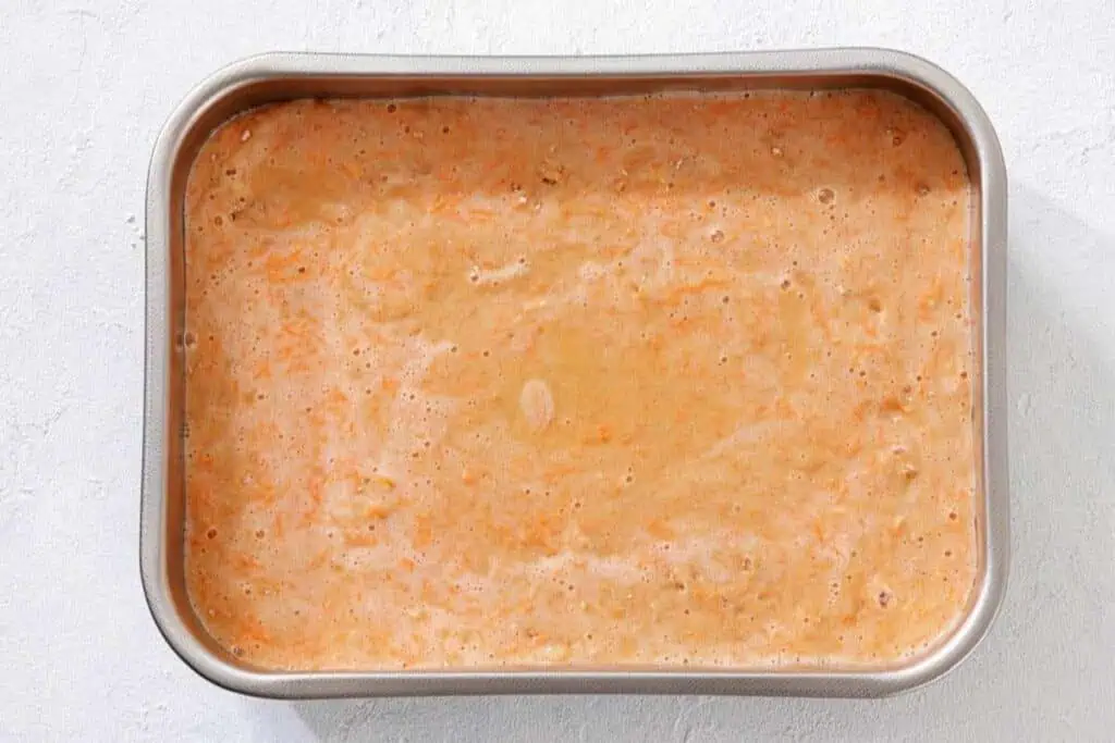 A rectangular metal baking pan filled with an unbaked, orange-speckled batter, sitting on a white surface.