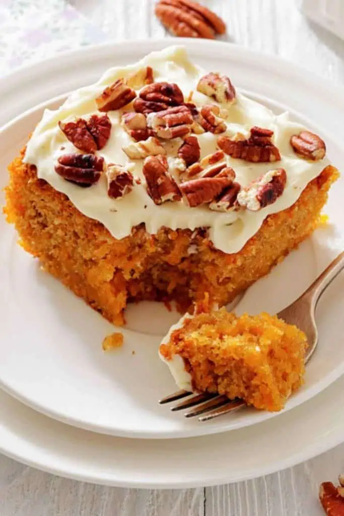 A slice of carrot cake with cream cheese frosting and chopped pecans on a white plate, with a fork holding a bite-sized piece.