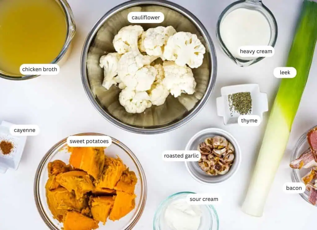 An overhead view of ingredients for cauliflower and sweet potato soup, including cauliflower, sweet potatoes, chicken broth, heavy cream, leek, bacon, roasted garlic, sour cream, thyme, and cayenne.