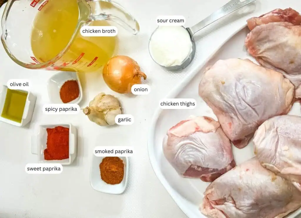 Ingredients for a recipe are displayed: chicken thighs, chicken broth, sour cream, an onion, garlic, olive oil, sweet paprika, spicy paprika, and smoked paprika.