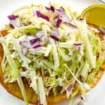 A tostada topped with shredded lettuce, cheese, and chopped red onion on a white plate with a lime wedge on the side.