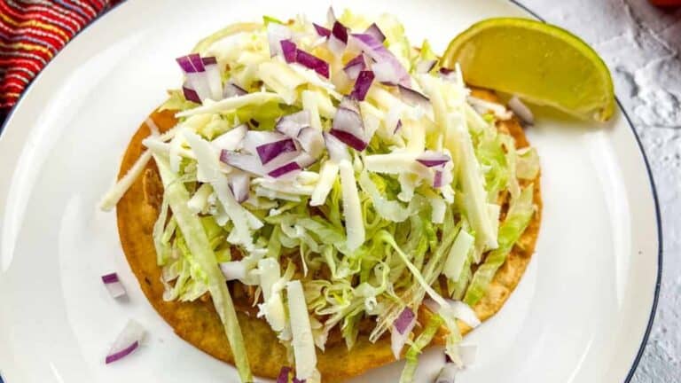 A tostada topped with shredded lettuce, cheese, and chopped red onion on a white plate with a lime wedge on the side.
