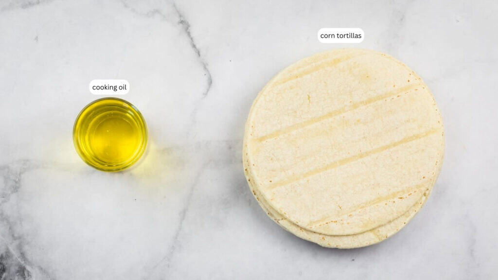 A small glass container of cooking oil sits next to a stack of corn tortillas on a light marble surface.