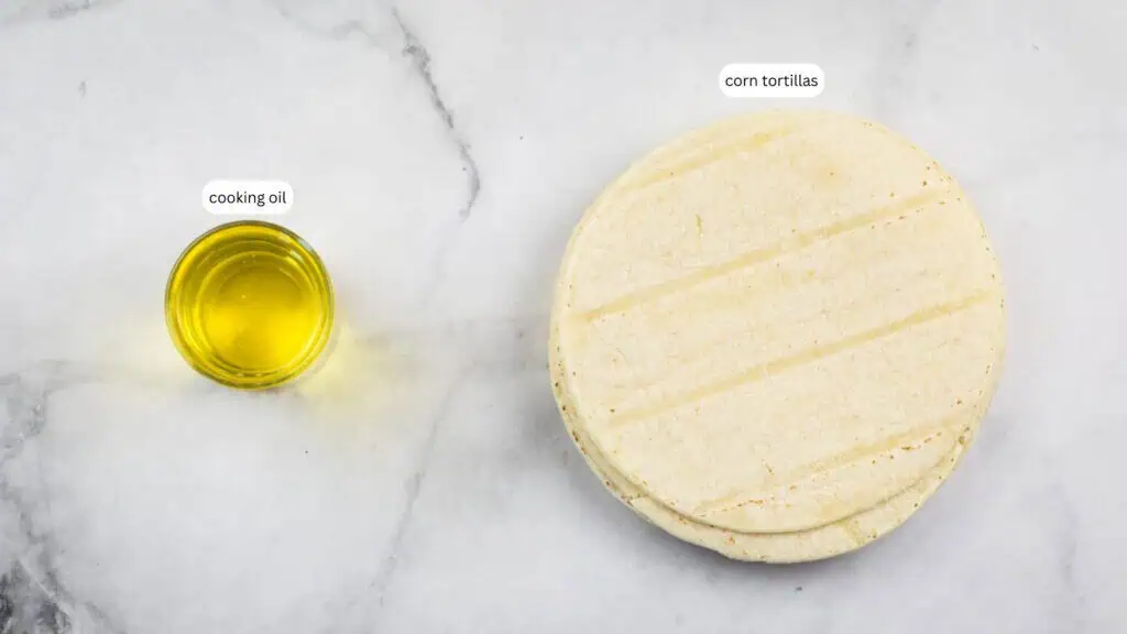 A small glass container of cooking oil sits next to a stack of corn tortillas on a light marble surface.