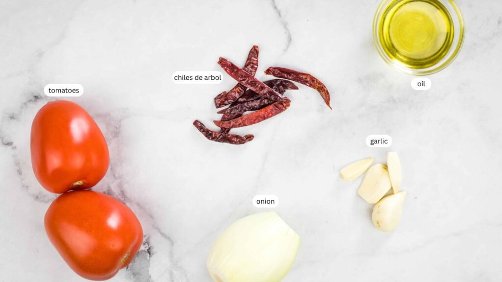 Two tomatoes, dried chiles de arbol, a small onion, garlic cloves, and a small dish of oil are arranged on a white marble surface. Labels identify each ingredient.