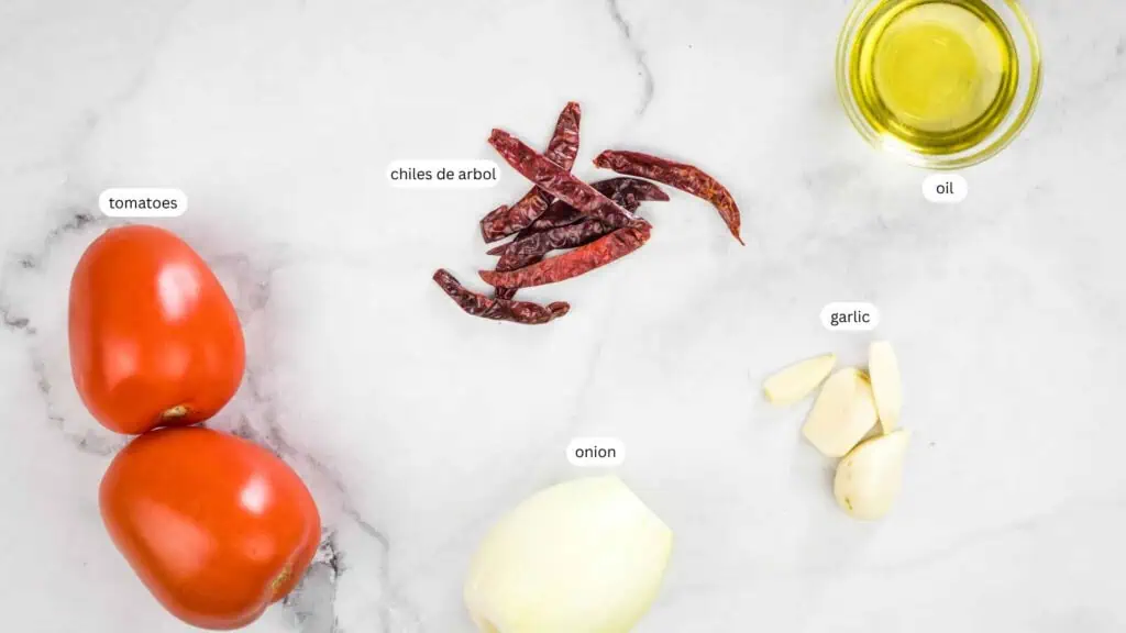 Two tomatoes, dried chiles de arbol, a small onion, garlic cloves, and a small dish of oil are arranged on a white marble surface. Labels identify each ingredient.