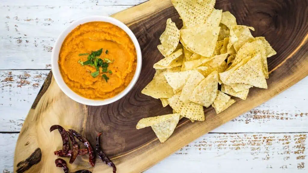 A bowl of orange dip garnished with herbs, served with tortilla chips and dried red chilies on a wooden board.