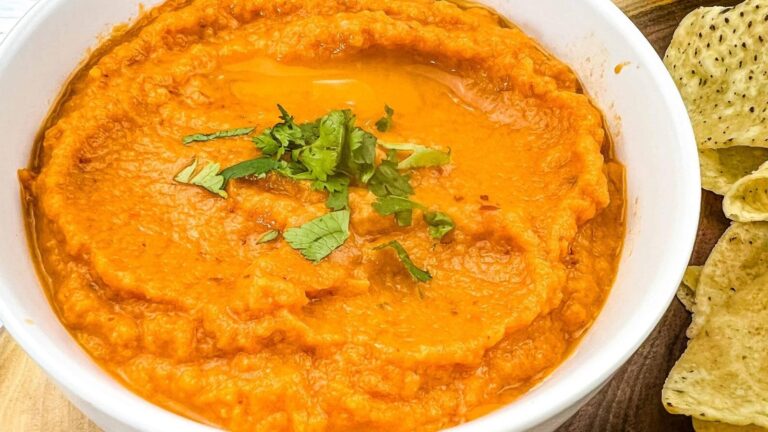 A white bowl filled with creamy orange dip, garnished with chopped cilantro, is placed next to a pile of tortilla chips on a wooden surface.
