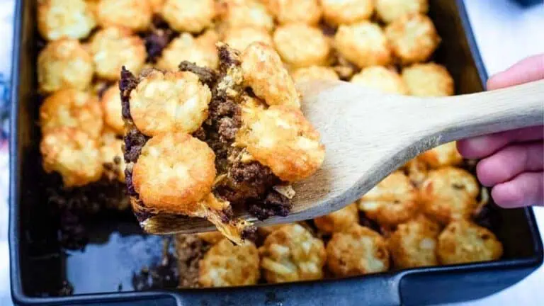 A wooden spatula lifts a serving of tater tot casserole with ground beef from a baking dish filled with more casserole.
