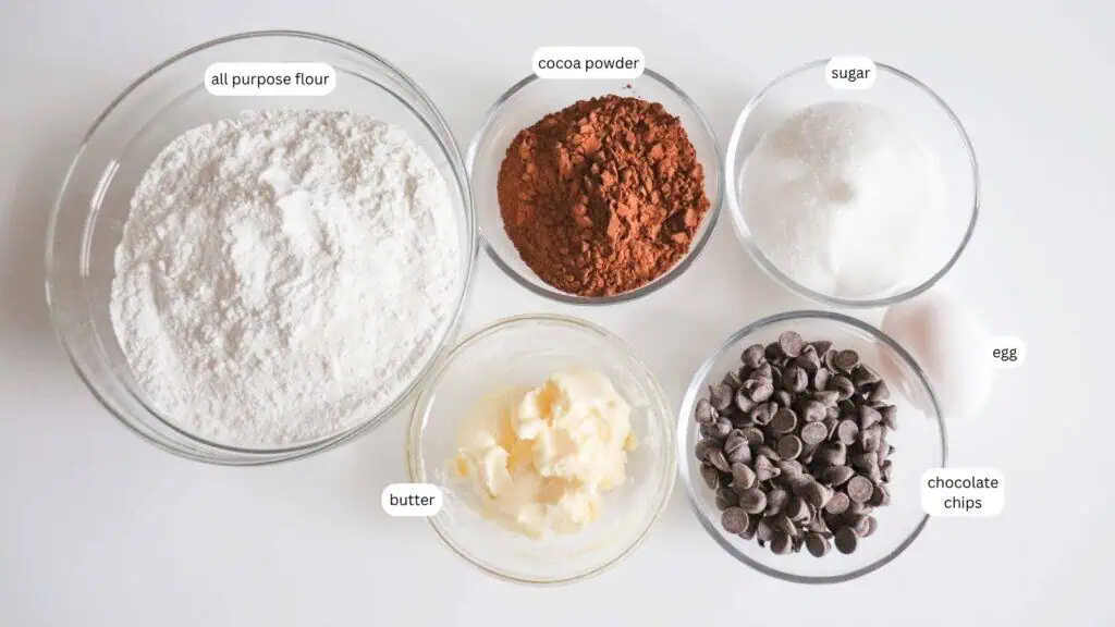 Five bowls containing all purpose flour, cocoa powder, sugar, butter, and chocolate chips, with an egg next to them, all labeled on a white surface.