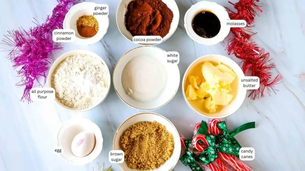 Top-down view of measured baking ingredients in bowls labeled: cinnamon powder, ginger powder, cocoa powder, molasses, unsalted butter, white sugar, brown sugar, egg, all-purpose flour, and candy canes.
