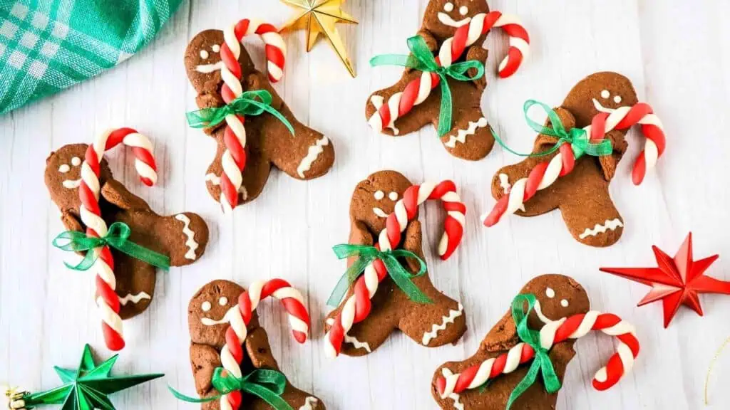Gingerbread cookies shaped like people, each holding a candy cane and decorated with green ribbon bows, are arranged on a white surface with red and gold star ornaments.