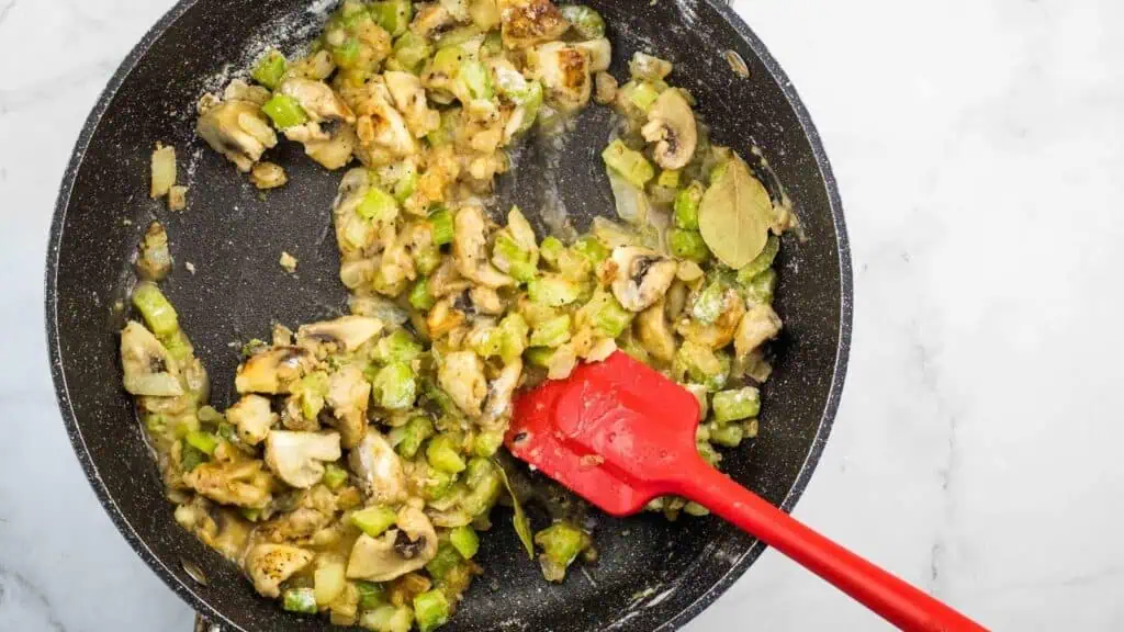 Sautéed mushrooms, celery, onions, and seasonings cooking in a black skillet with a red spatula on a white surface.
