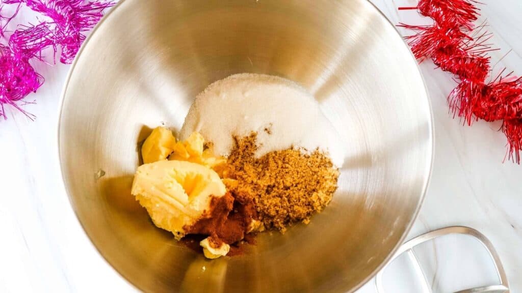A mixing bowl containing butter, brown sugar, white sugar, and spices on a white surface with pink and red tinsel decorations nearby.