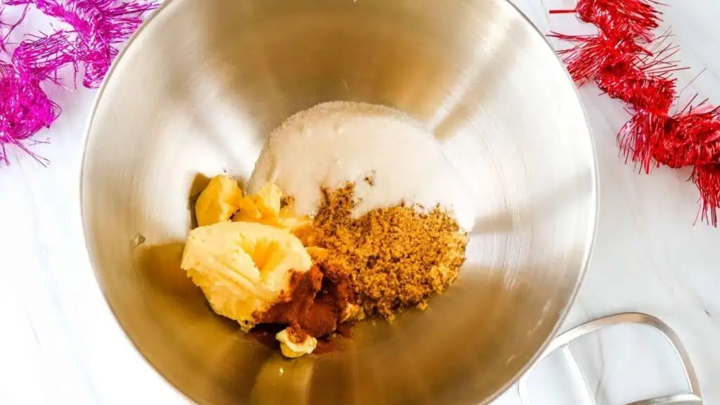 A mixing bowl containing butter, brown sugar, white sugar, and spices on a white surface with pink and red tinsel decorations nearby.