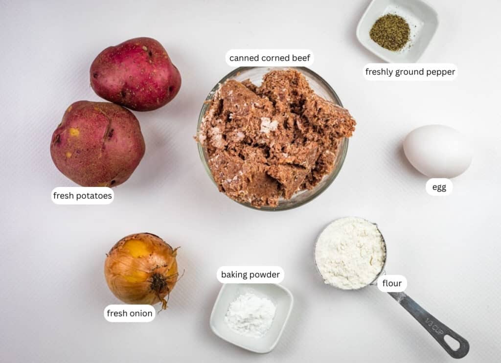 Top-down view of ingredients for crispy corned beef fritters, including fresh potatoes, fresh onion, canned corned beef, baking powder, flour, an egg, and freshly ground pepper, all labeled.