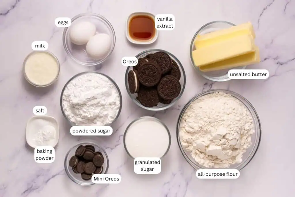 Bowls of baking ingredients on a marble surface, including eggs, milk, salt, baking powder, powdered sugar, Mini Oreos, Oreos, granulated sugar, vanilla extract, unsalted butter, and all-purpose flour.
