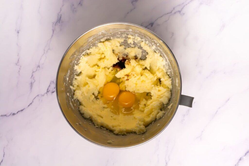 A metal mixing bowl containing creamed butter, sugar, two raw eggs, and vanilla extract on a marble countertop.