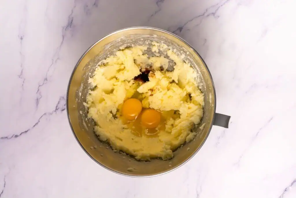 A metal mixing bowl containing creamed butter, sugar, two raw eggs, and vanilla extract on a marble countertop.