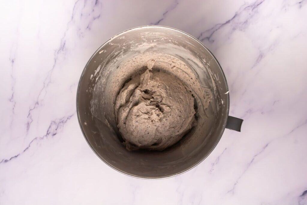 A metal mixing bowl filled with light brown bread dough sits on a white marble surface.