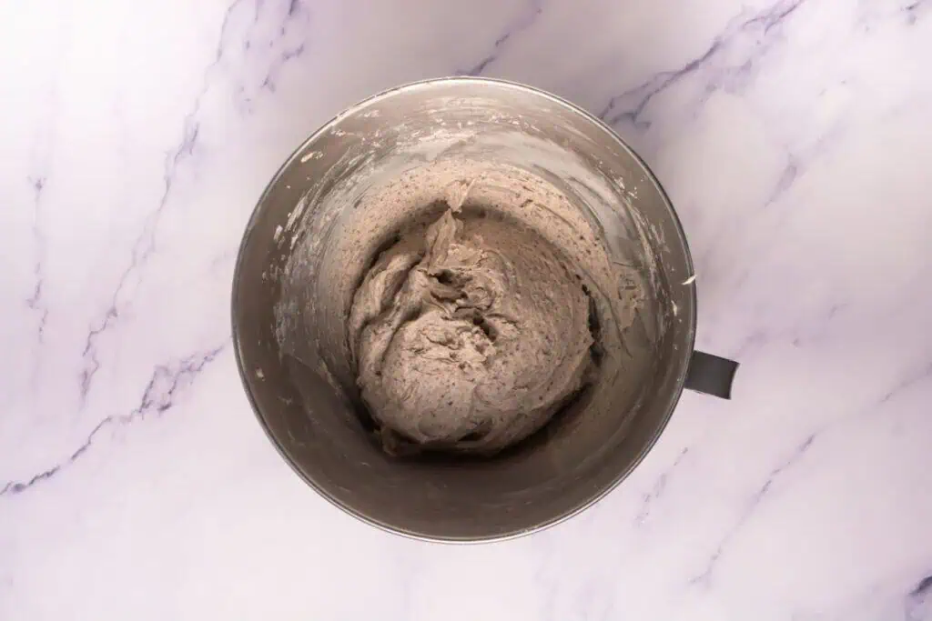 A metal mixing bowl filled with light brown bread dough sits on a white marble surface.