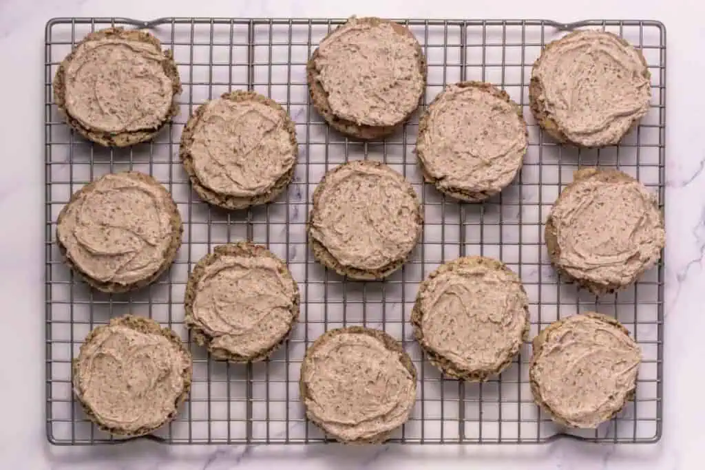 Round cookies with light brown frosting arranged in rows on a wire cooling rack over a white marble surface.