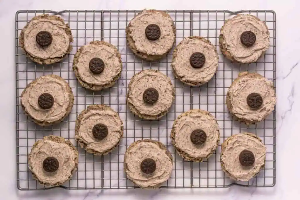 A cooling rack holds 14 round cookies topped with light brown frosting and a chocolate cookie piece in the center of each.